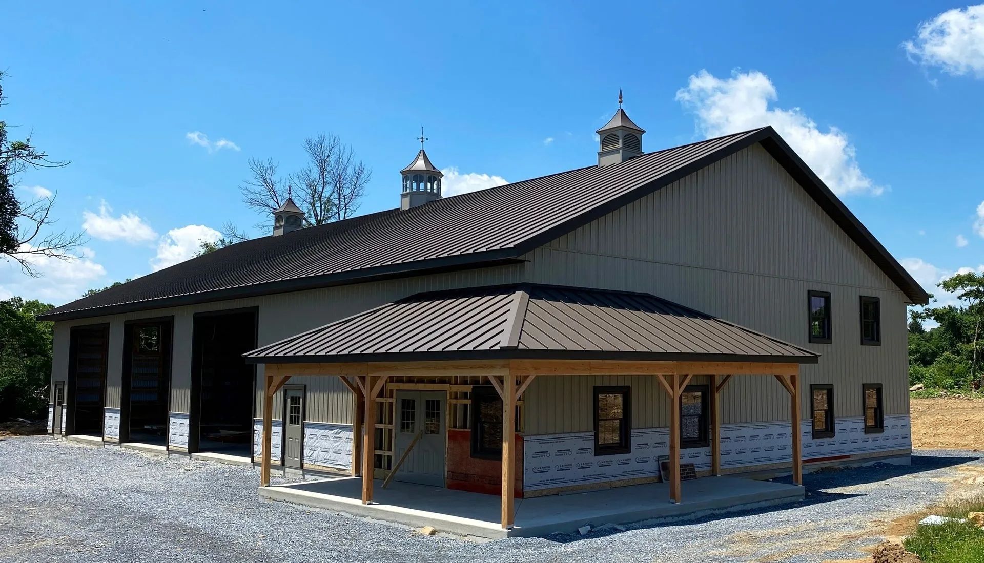 Large tan barn with brown roof and a porch, set on a gravel lot under a blue sky.