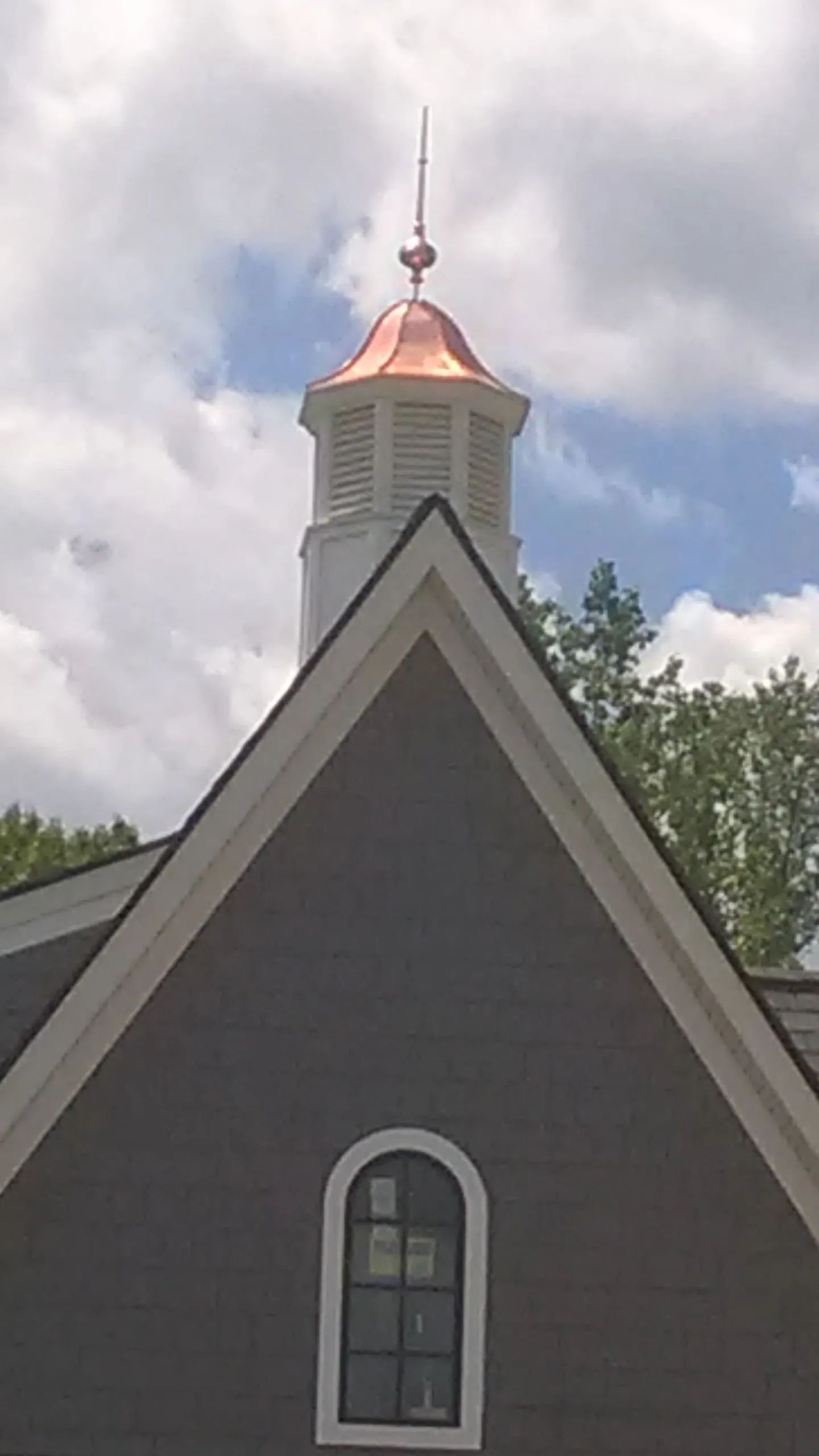 Copper-topped cupola with lightning rod atop a gray building with a triangular roof and arched window against a cloudy sky.