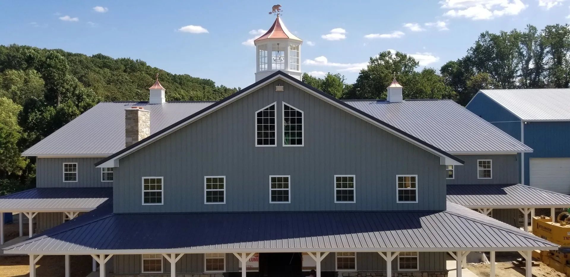 A large gray barn with a blue roof, white trim, and a cupola. Green trees in the background under a blue sky.