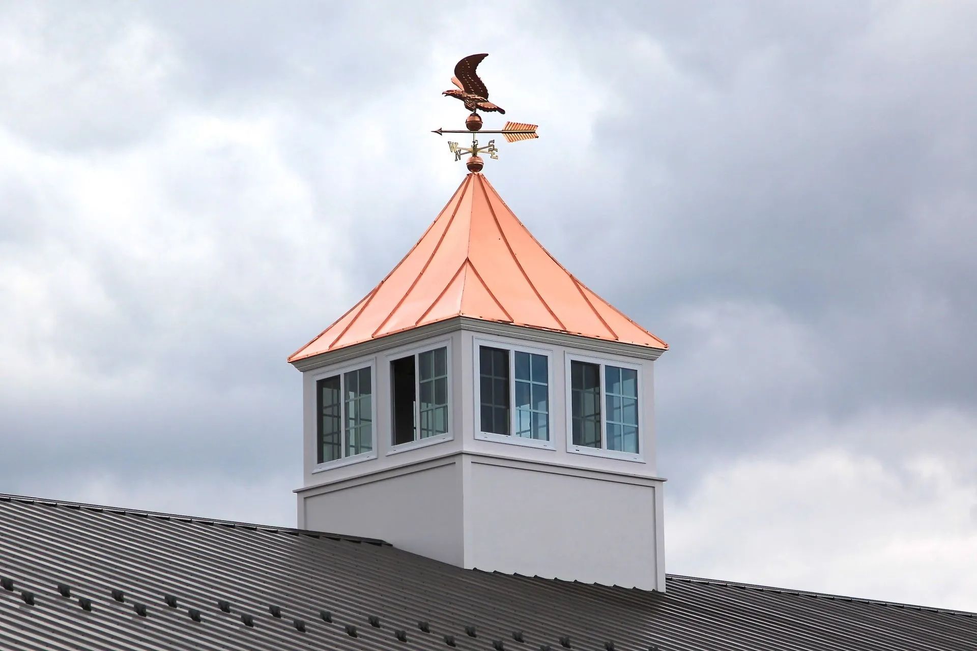 Copper-roofed cupola with an eagle weathervane atop a building, against a cloudy sky.