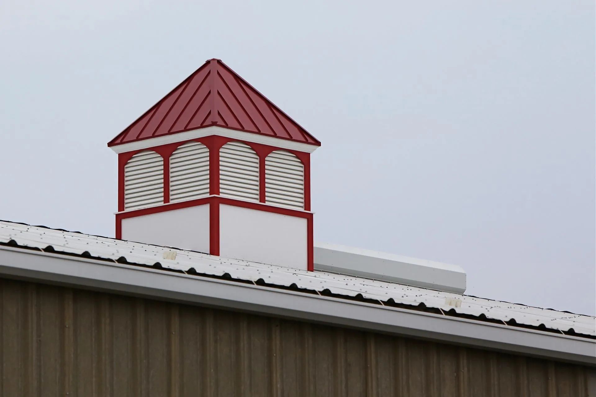 Red-roofed cupola on a white and red base atop a tan building, against a gray sky.