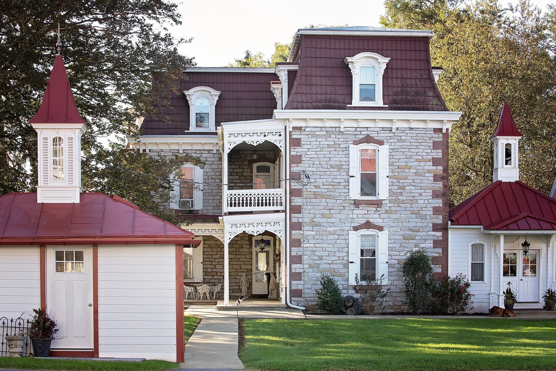 Stone mansion with a red roof and white trim, flanked by matching white outbuildings.