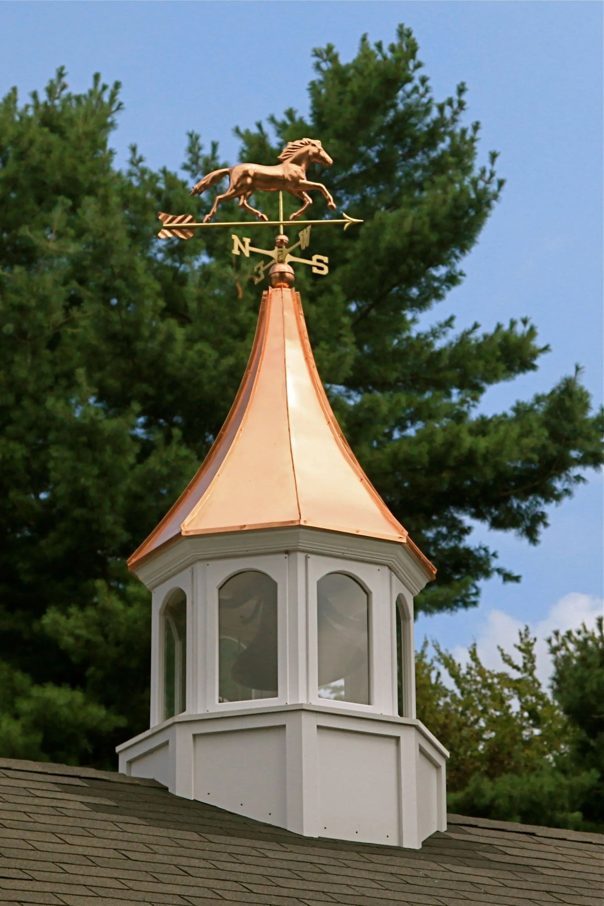 Copper horse weathervane atop a white, windowed cupola on a roof, with trees in the background.