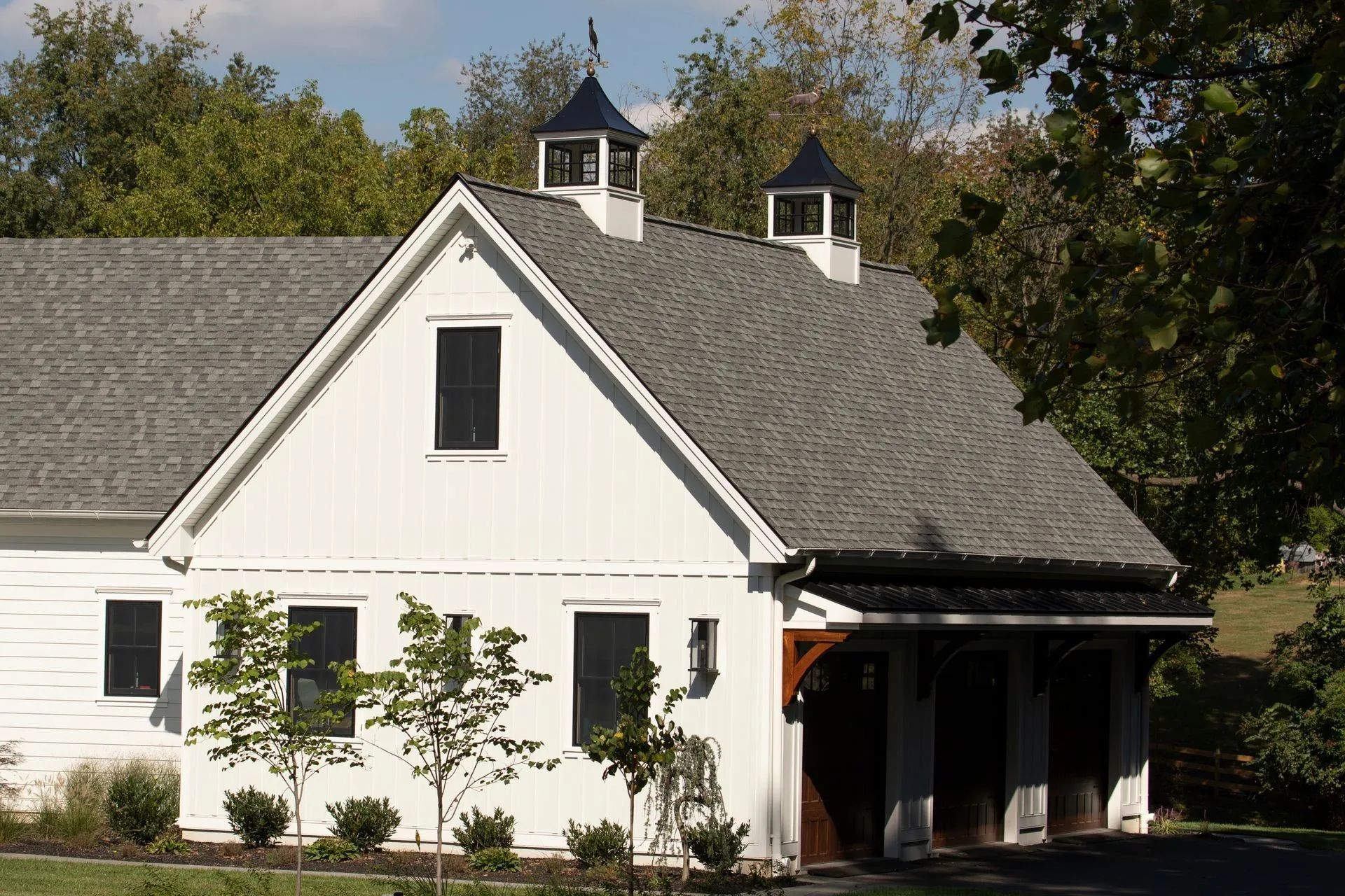 White barn with gray roof, black shutters, and cupolas. Trees in the background, sunny day.