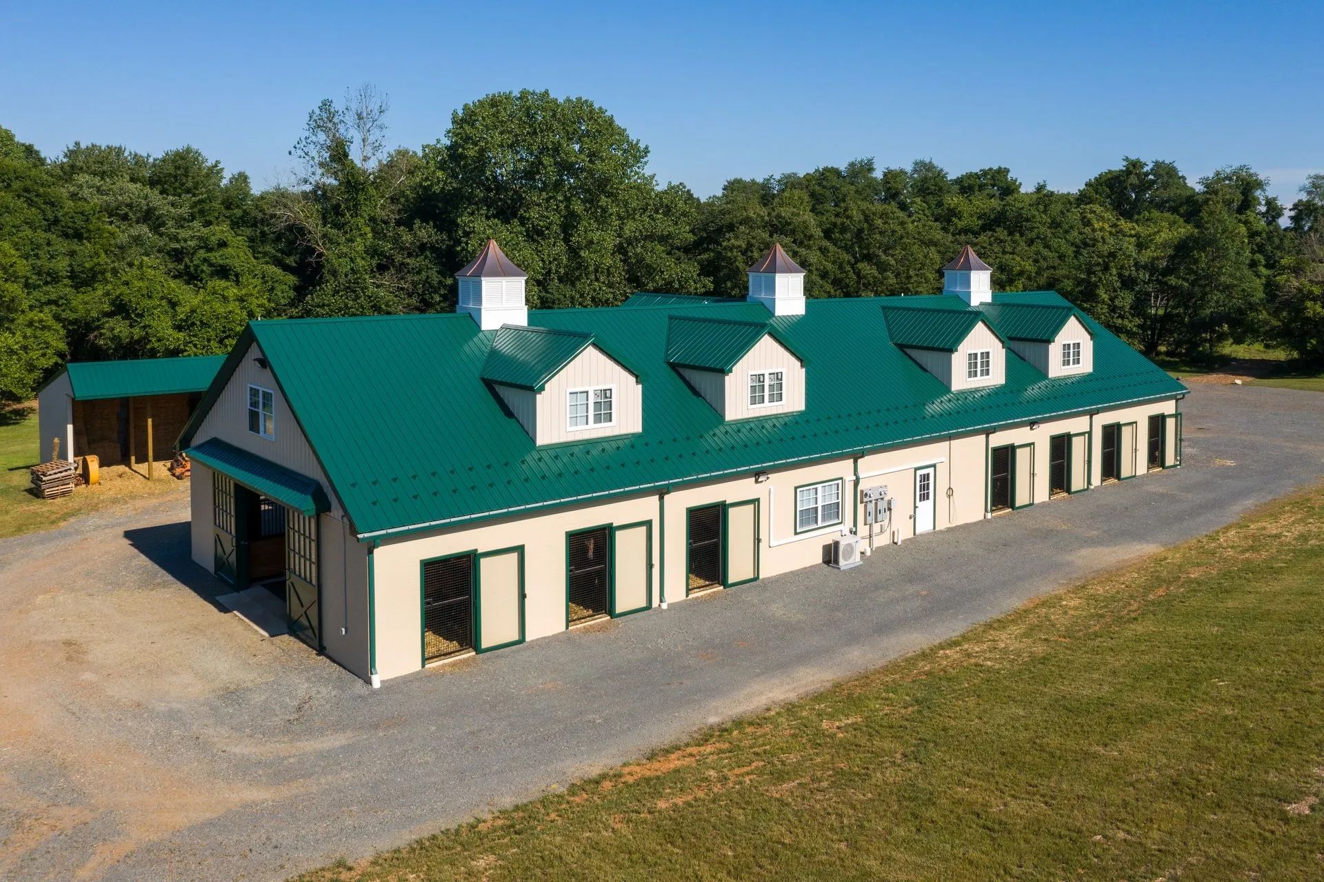 A long, beige horse stable with a green roof, dormers, and open stalls; exterior view.