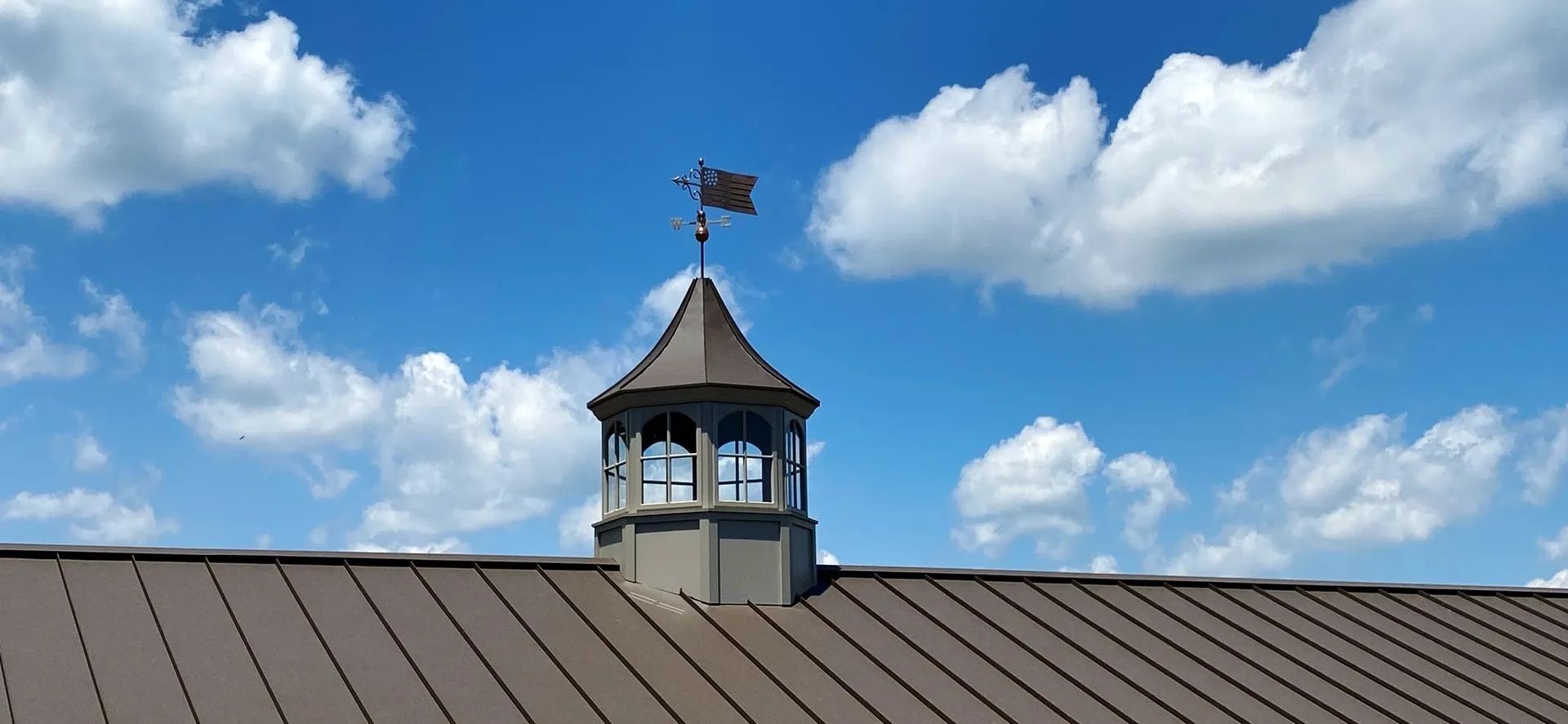 A brown roof with a cupola and a weather vane against a blue sky with fluffy white clouds.