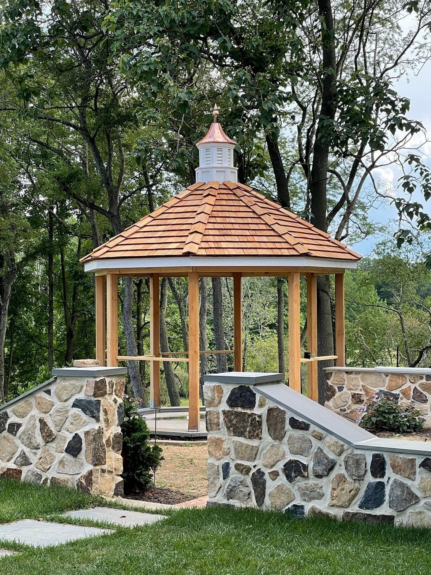 Wooden gazebo with cedar shingle roof, on stone wall, in a garden setting, with a copper finial.