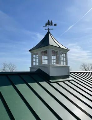 Green metal roof with white cupola and weather vane against a blue sky.