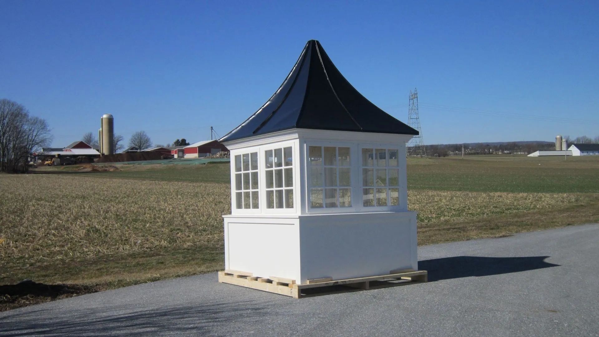 White gazebo with dark blue roof, on a pallet, sits on a road with fields and a farm in the background.
