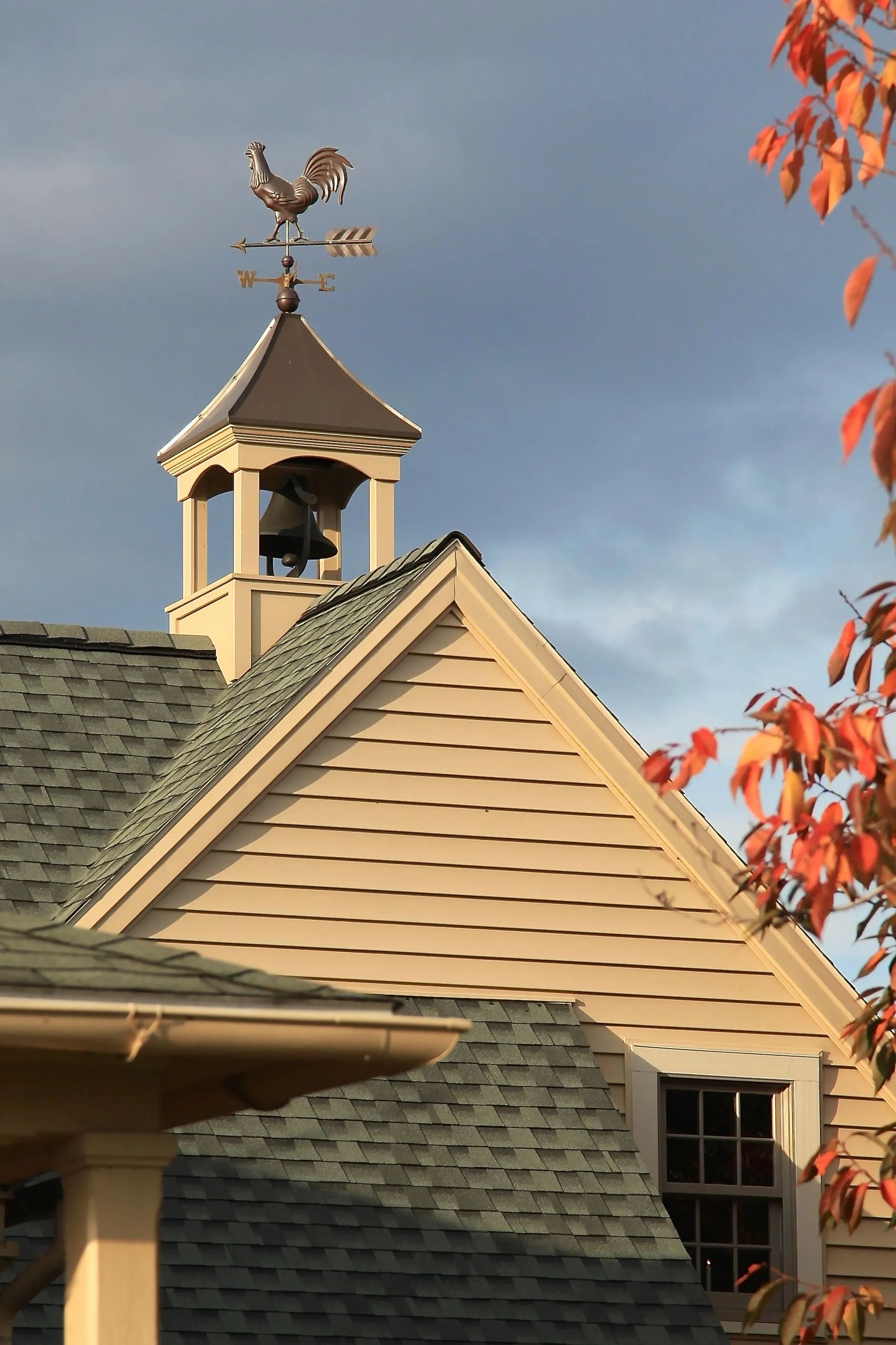 Tan building with green roof, bell tower, and weather vane against a blue sky, with autumn foliage.