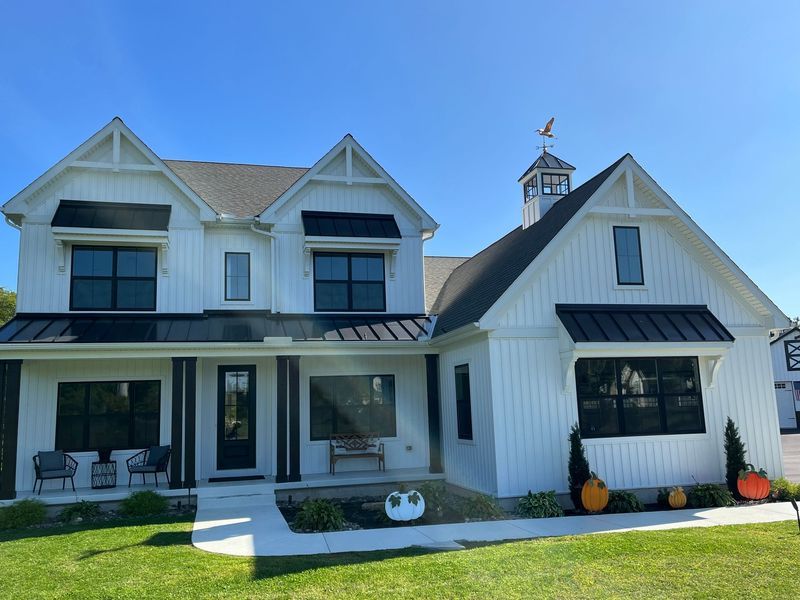 White farmhouse with black trim, a cupola, and pumpkins on the lawn under a bright blue sky.