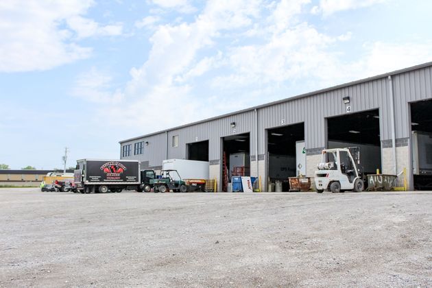 Truck loading bays at a gray warehouse with forklifts and parked delivery trucks on a gravel lot