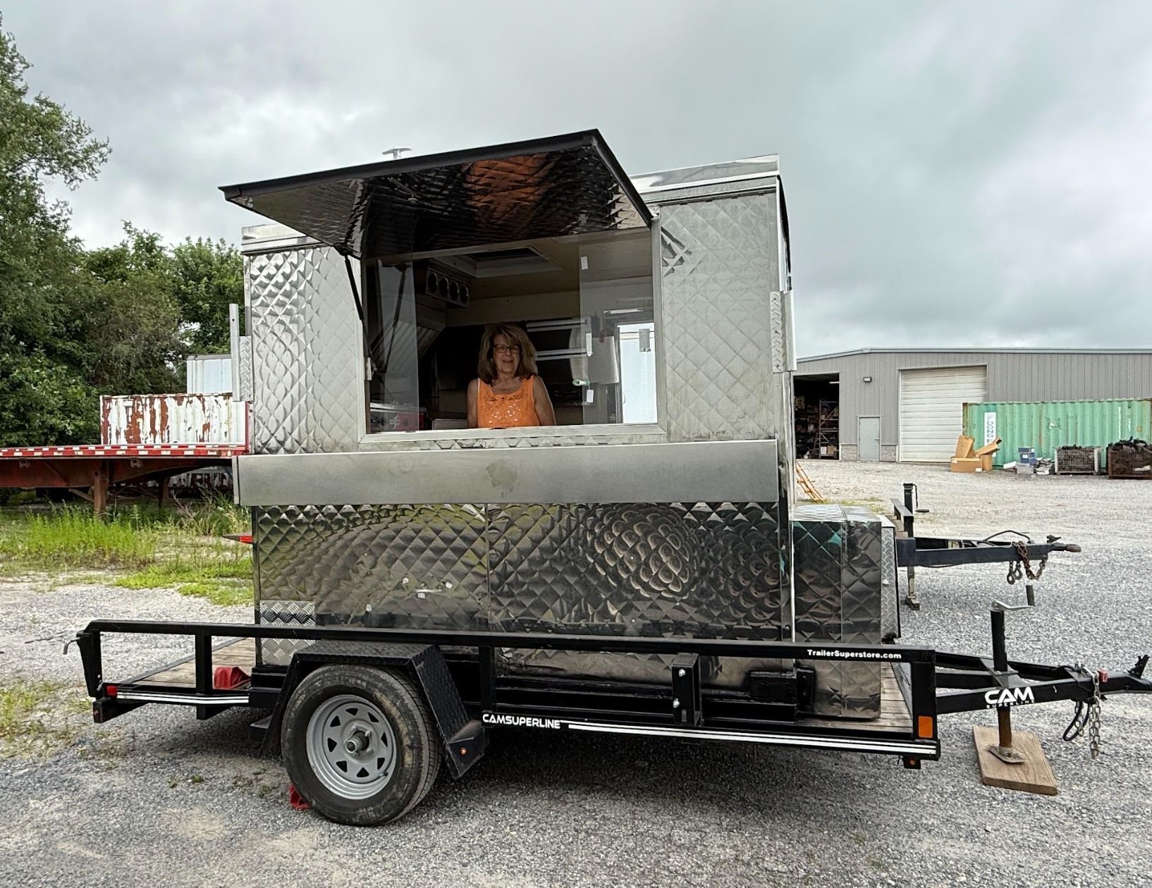Silver food trailer with open serving window and person inside, parked on gravel under cloudy sky