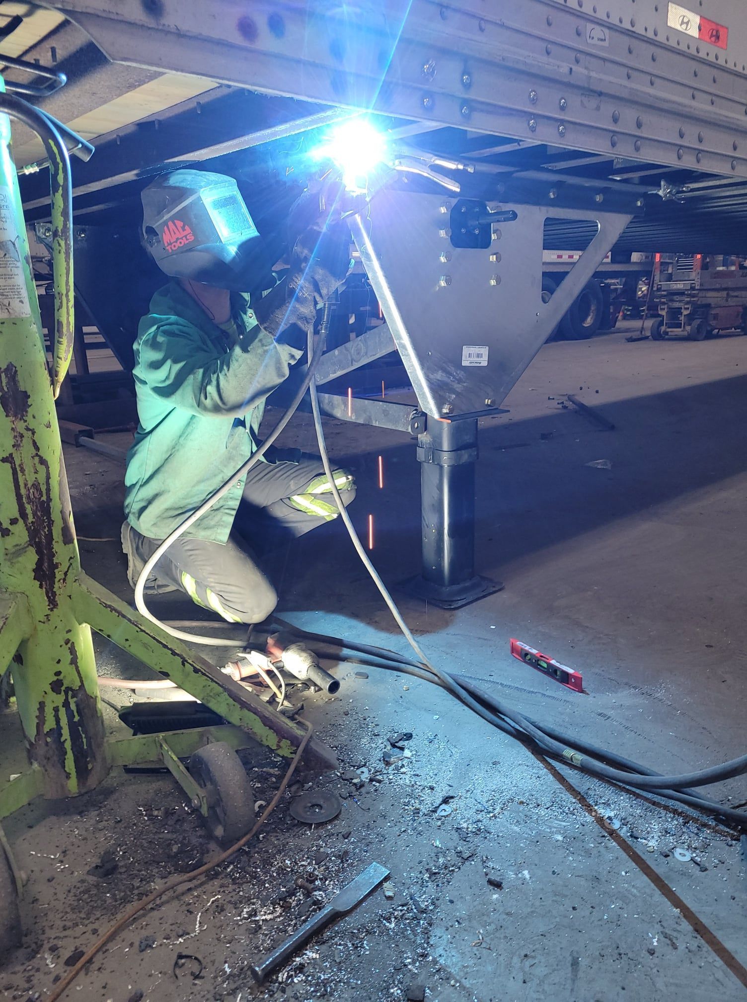 Welder in green protective suit welding under a large metal structure, bright blue arc light at night