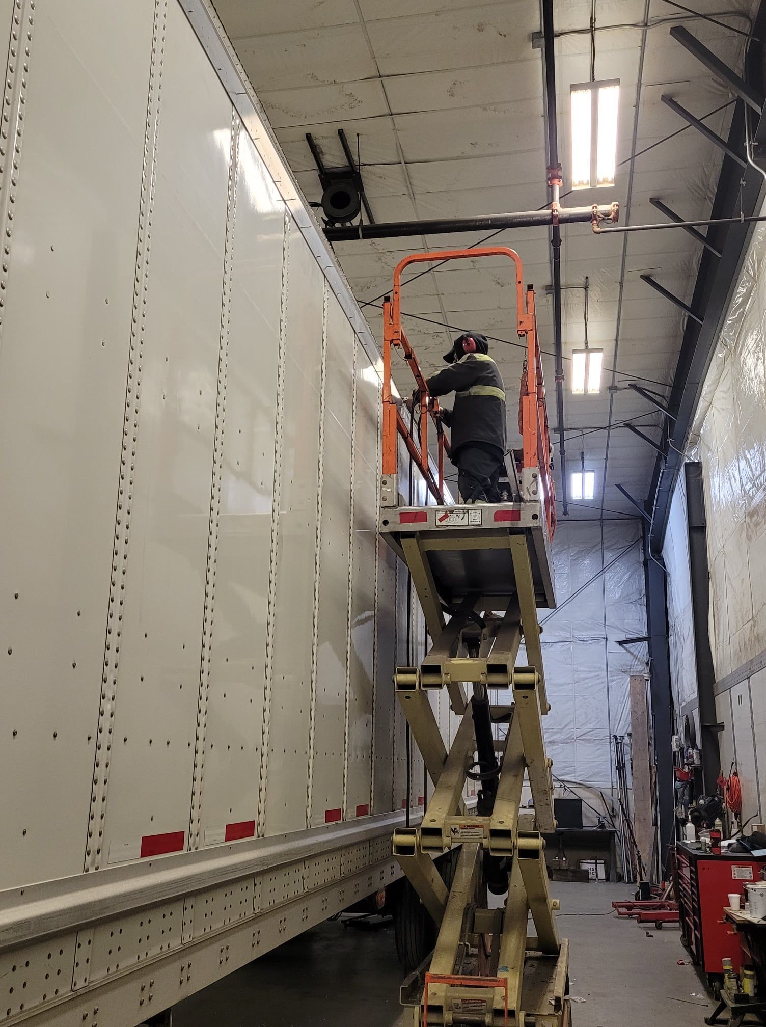 Worker on a scissor lift inspecting the side of a white semi-trailer in a warehouse.