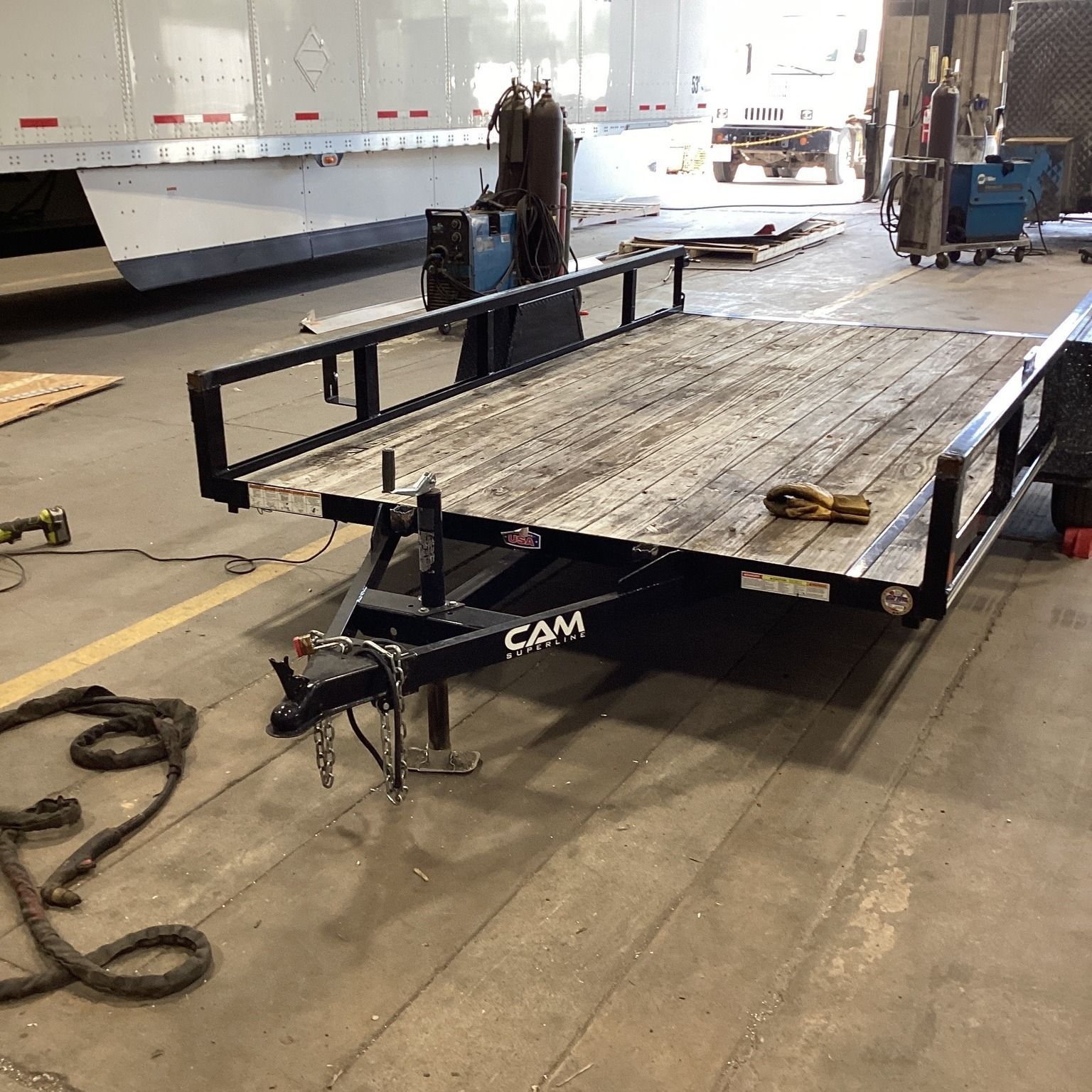 Flatbed trailer with a black ramp and chains inside an industrial warehouse, viewed from the rear.