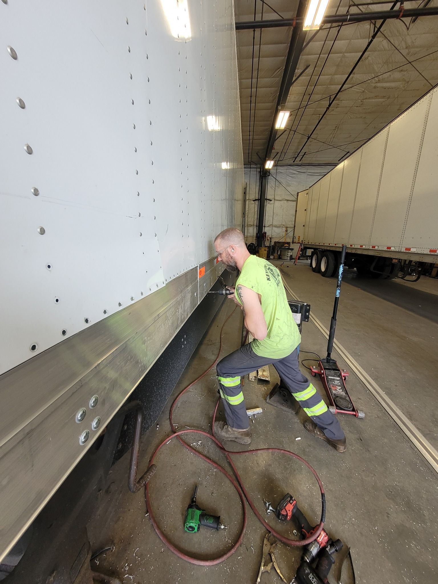 Worker in neon vest inspects a large white truck in a garage, with hoses coiled on the floor.