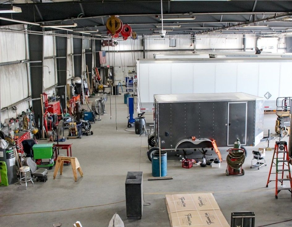 Large workshop interior with a gray trailer, tools, equipment, and ladders scattered around