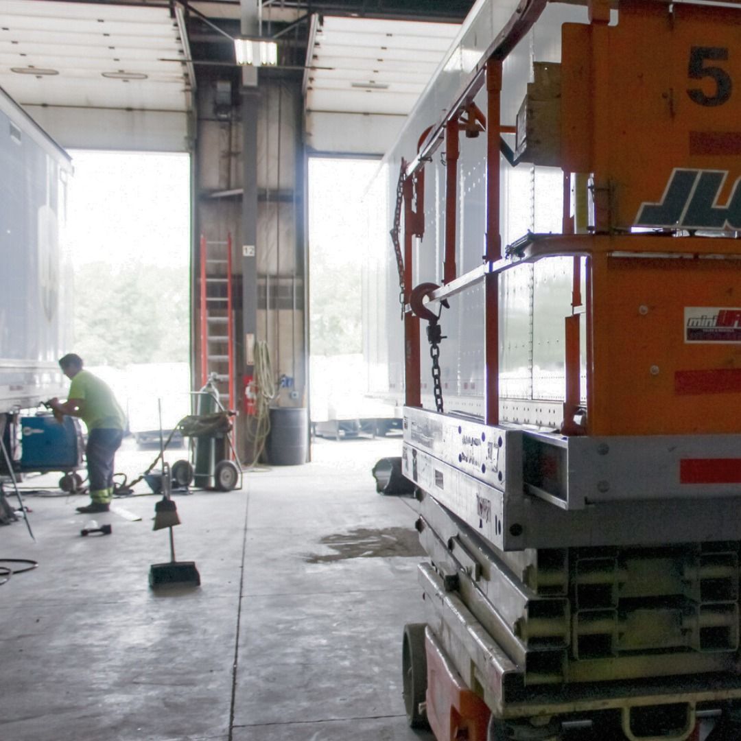 Worker in a warehouse beside a large orange scissor lift and cleaning tools near an open loading bay