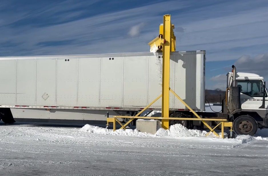Large white truck trailer beside yellow industrial equipment on snowy ground