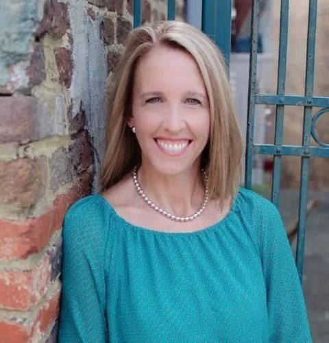 Smiling woman in a teal top and pearl necklace standing by a brick wall and metal gate