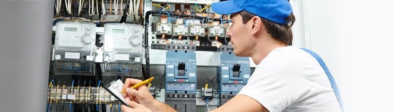 A man in a blue hat is writing on a clipboard in front of an electrical panel.