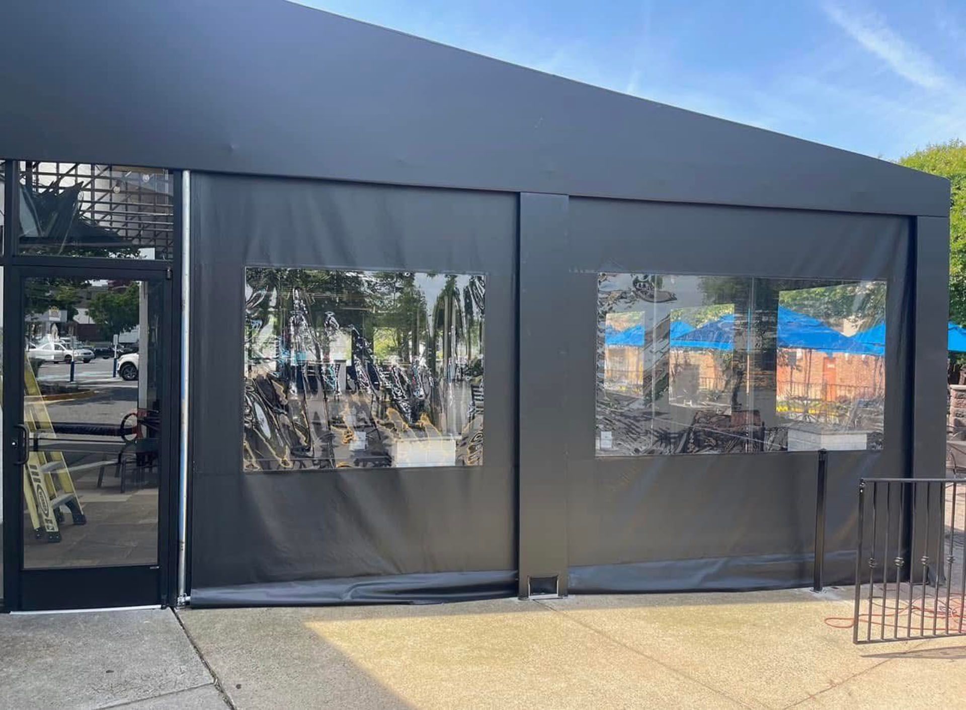 A black tent with a lot of windows is sitting on the sidewalk in front of a building.