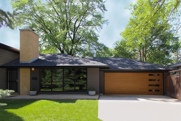 A modern one-story suburban house with a brick chimney, dark brown exterior, large front windows, and a wood garage door.