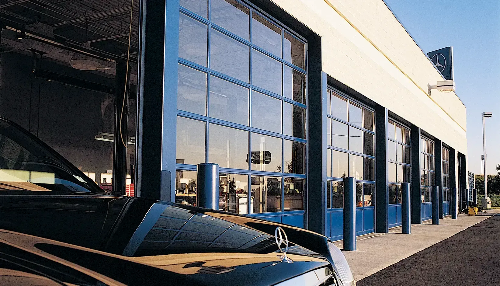 Car dealership exterior with a black car parked beside glass service bay doors on a sunny day