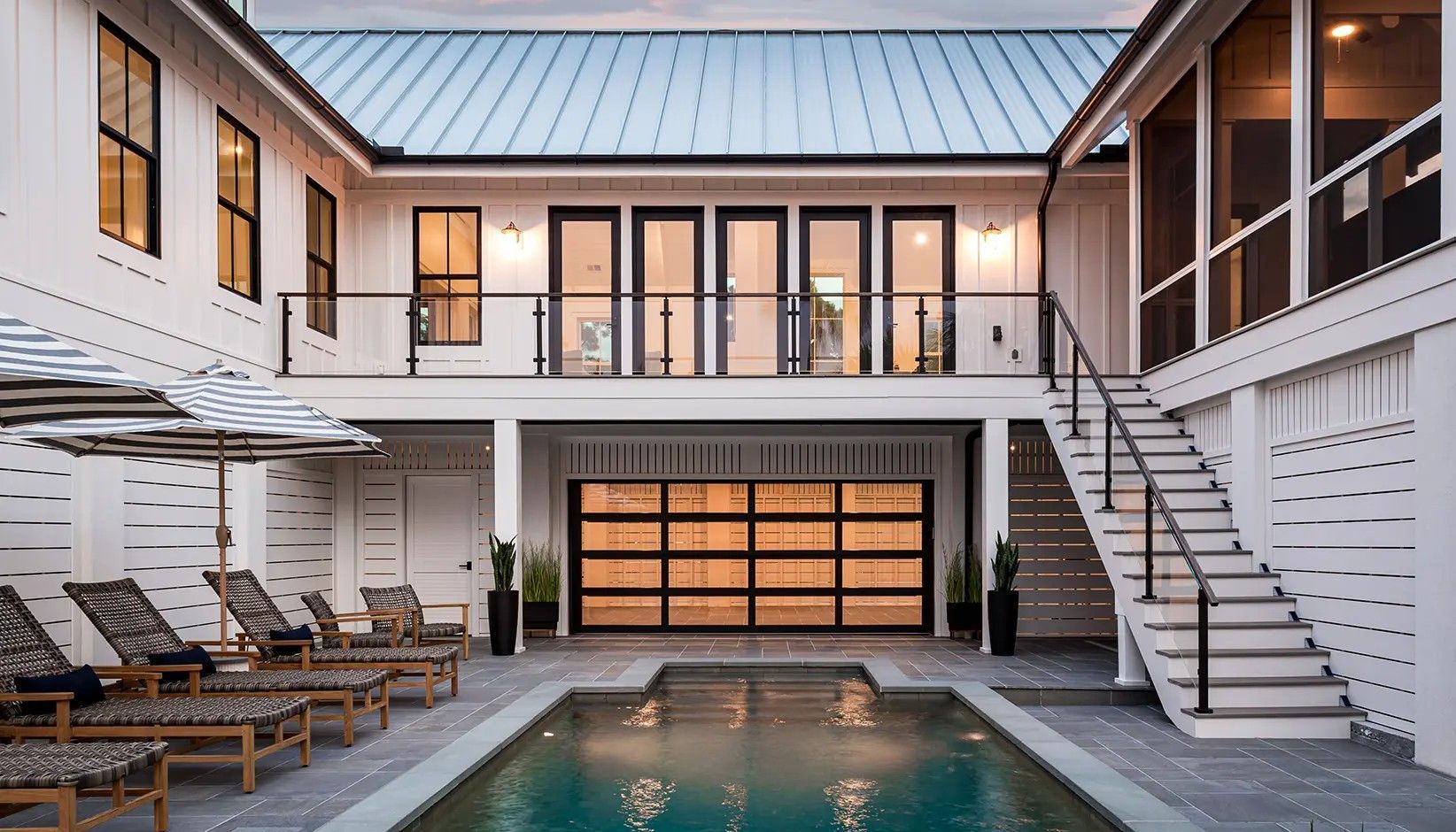 A modern white house with a metal roof, a pool, lounge chairs, a glass garage door, and stairs to an upper deck.