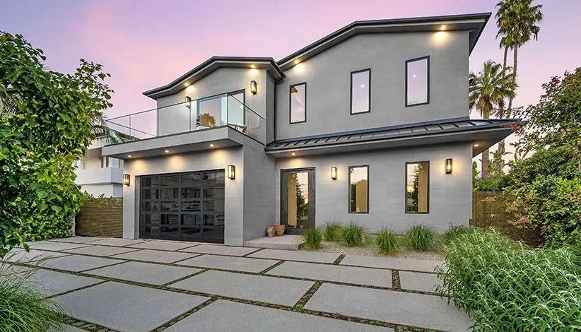 A two-story, grey modern house with a garage, glass balcony, and paved driveway at twilight.