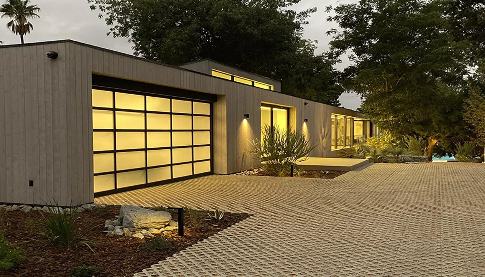 A modern, single-story home with a grid-patterned garage door, wood siding, and a paved driveway at dusk.