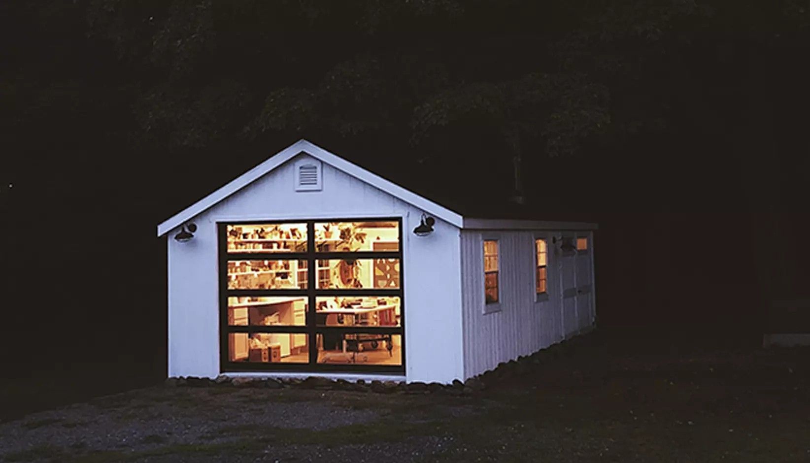 A small white detached garage or shed with a large, illuminated glass-paneled garage door at night.