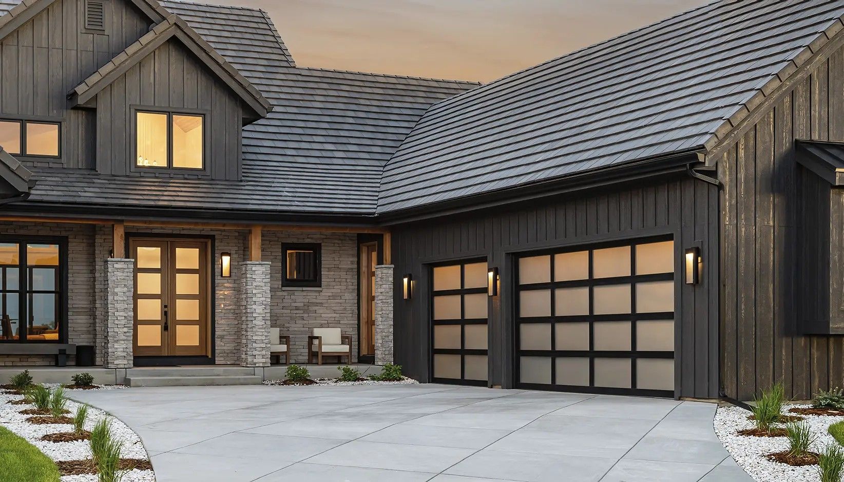 Modern suburban home with dark wood siding, stone accents, glass-panel garage doors, and a gray shingled roof at twilight.
