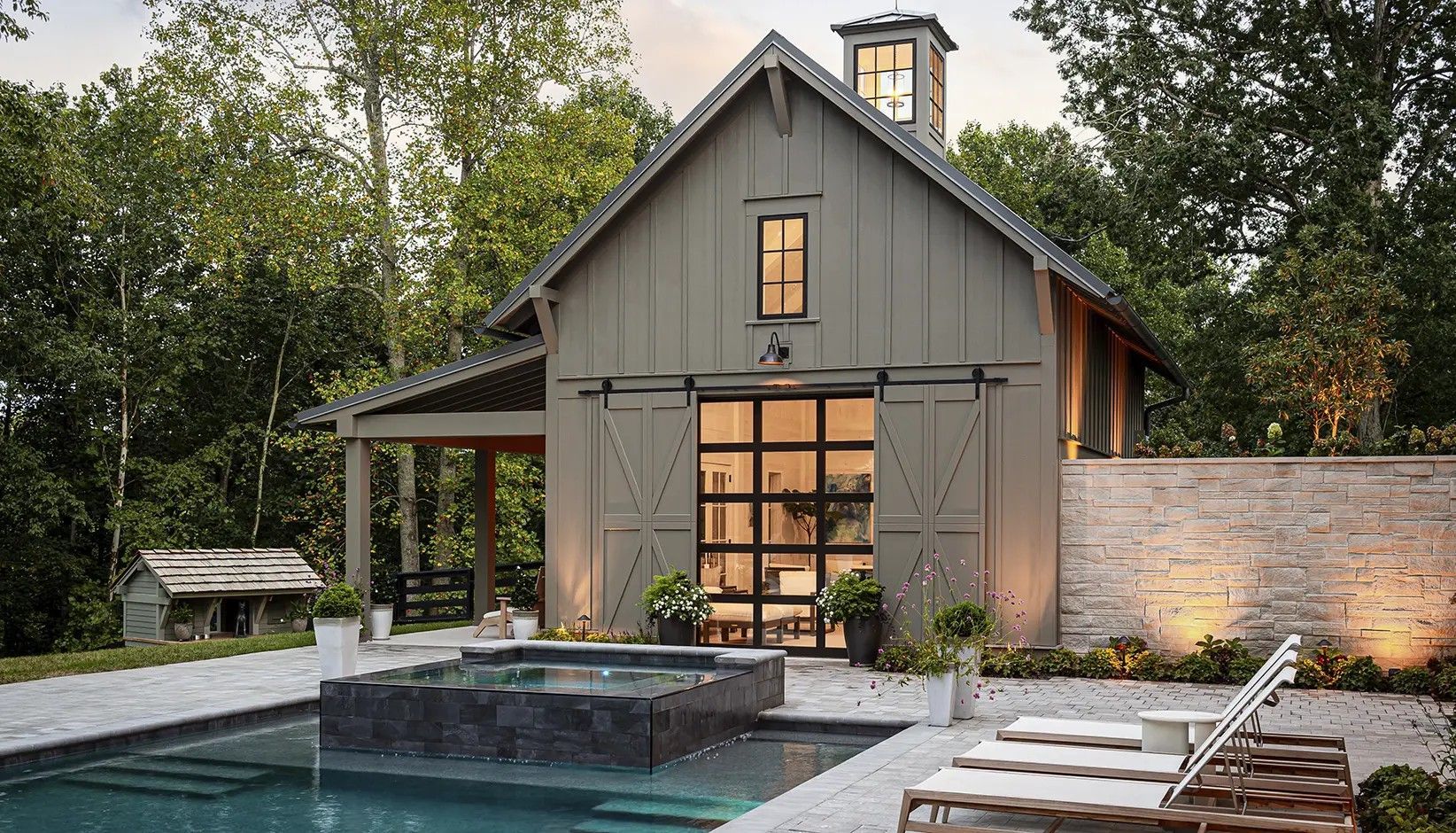A modern gray barn-style house with a glass garage door, a pool with a hot tub, and lounge chairs in a wooded setting.
