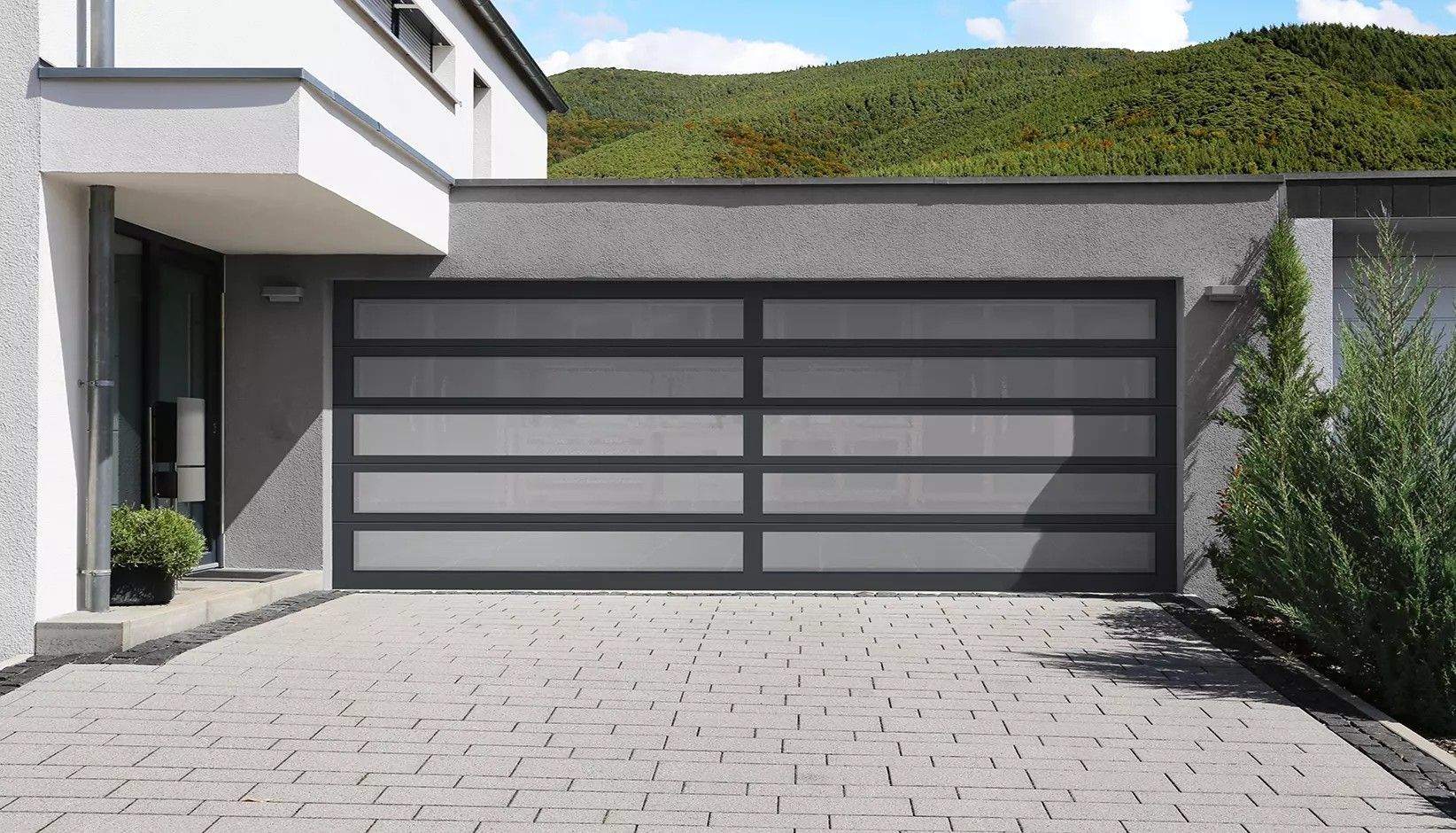 A modern garage door with grey panels and dark grey horizontal trim frames in front of a stone driveway and house.