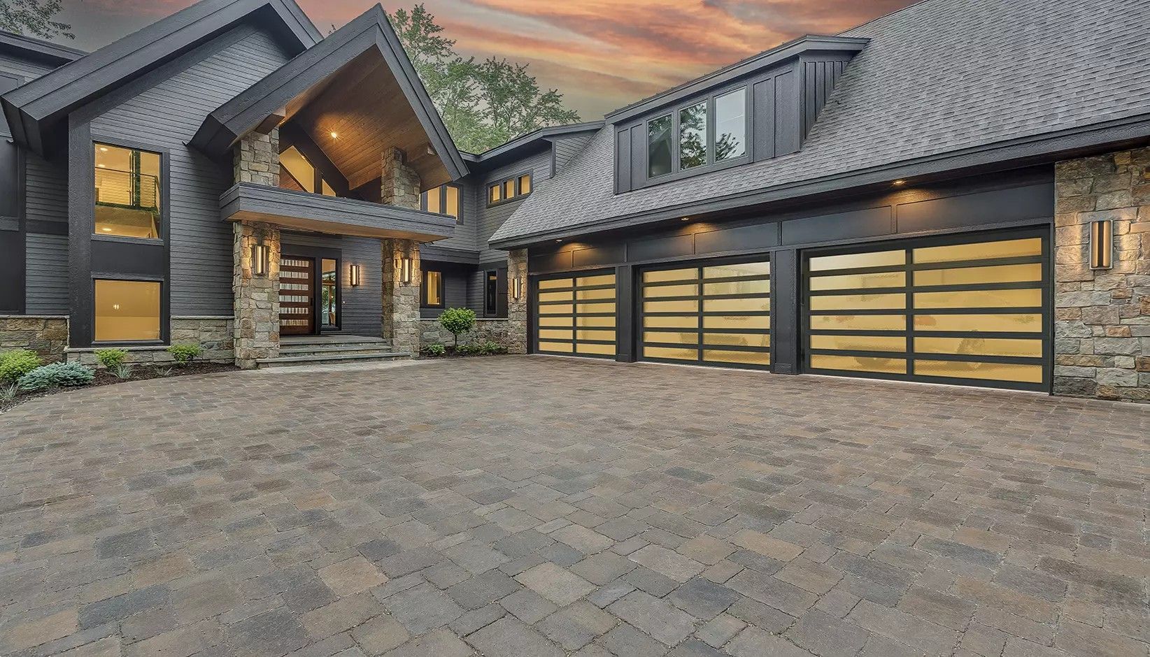 A modern home with dark siding, stone accents, and a triple-car garage, fronted by a large stone paver driveway.