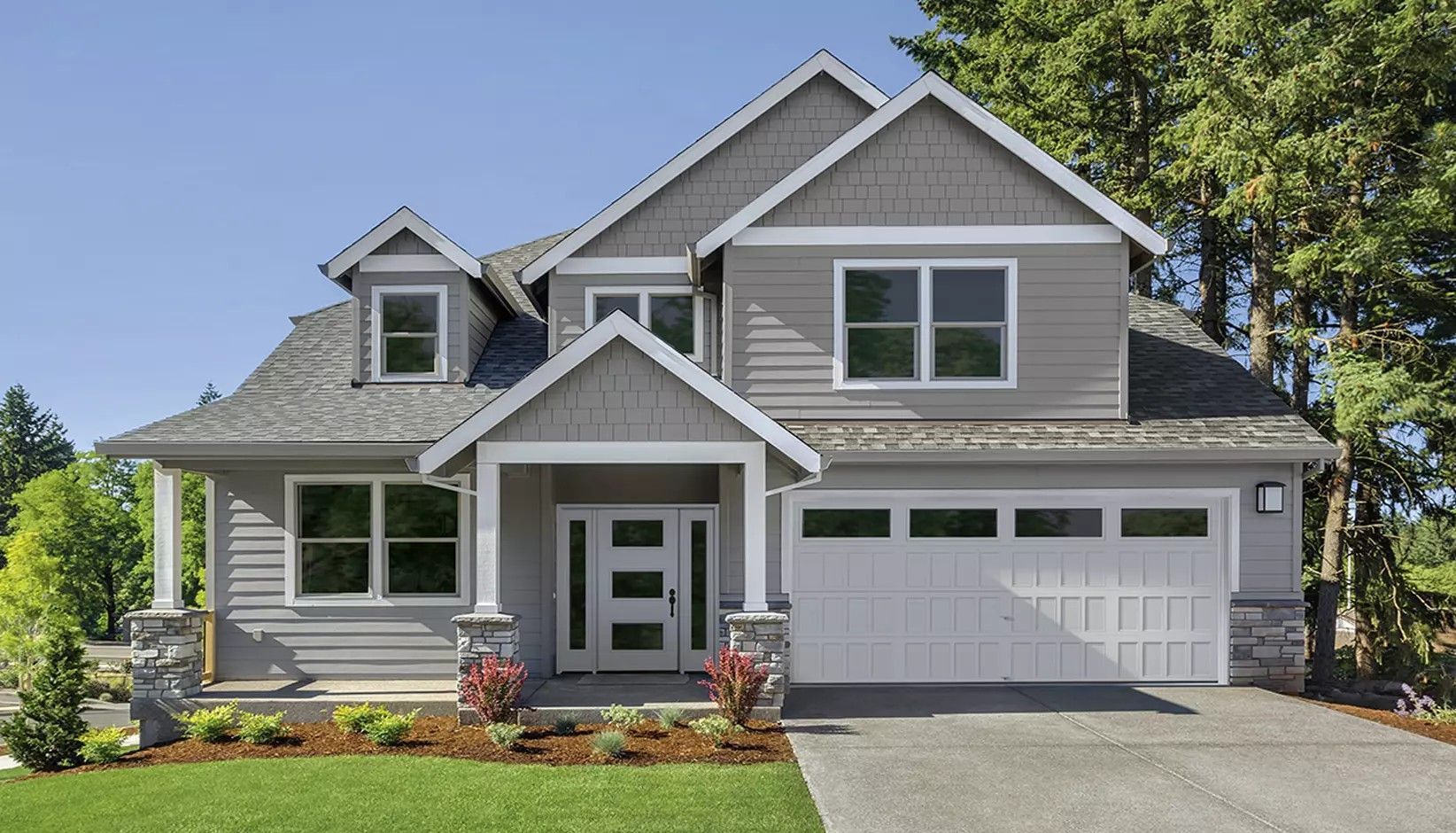 A two-story, gray-sided house with a white trim, a front porch, and a two-car garage, set against a blue sky and trees.