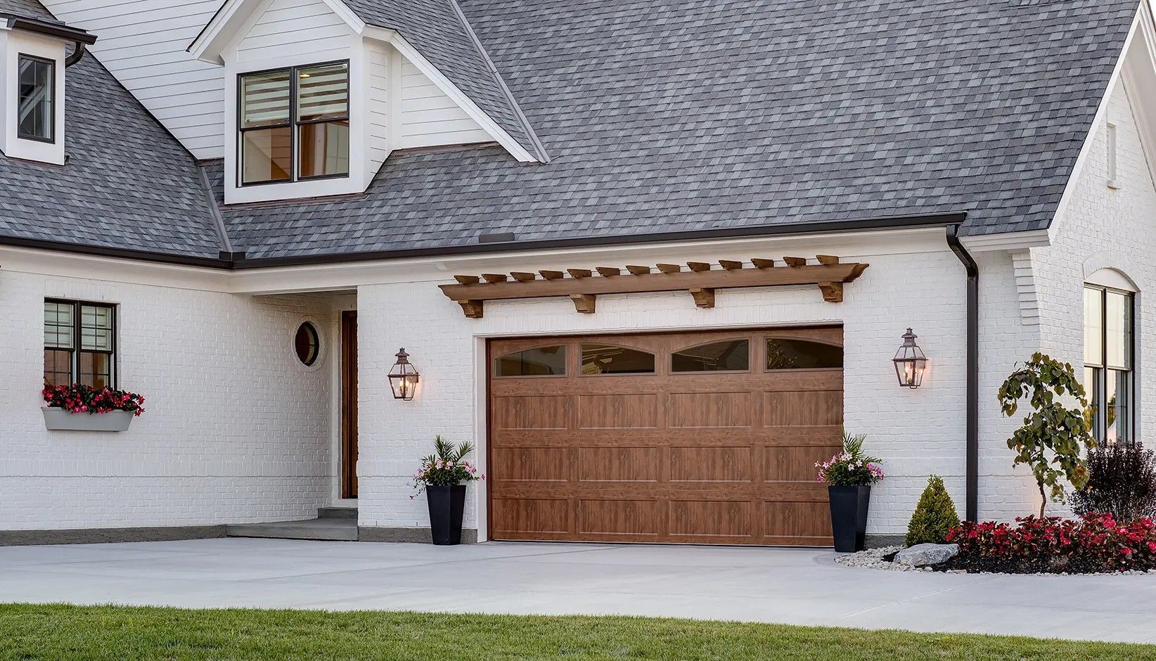 A white brick house with a dark wooden garage door, a wooden pergola, and a gray shingled roof.