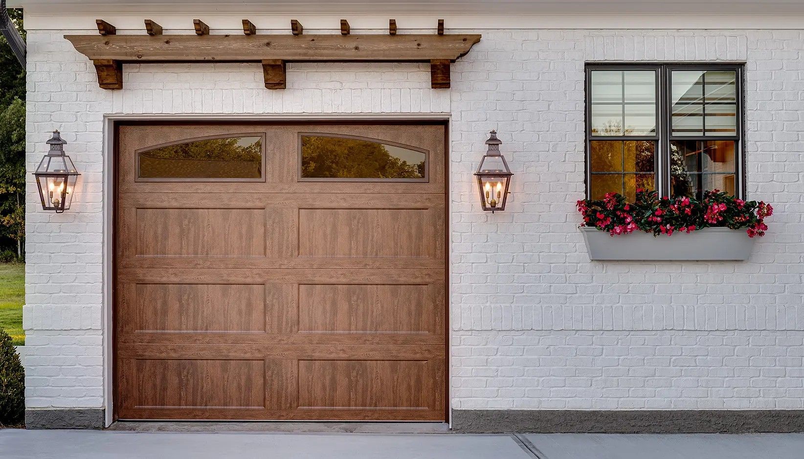 A wood-toned garage door with windows next to a white brick wall featuring a window box with flowers and two wall lamps.