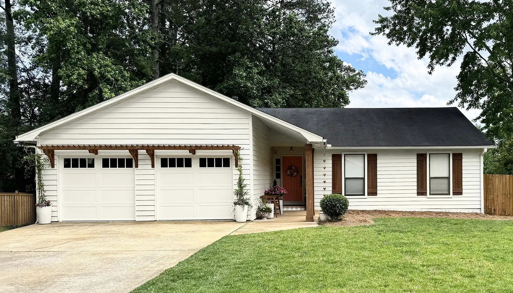 A white, single-story house with a two-car garage, brown shutters, and a front porch, set on a grassy lawn with trees.