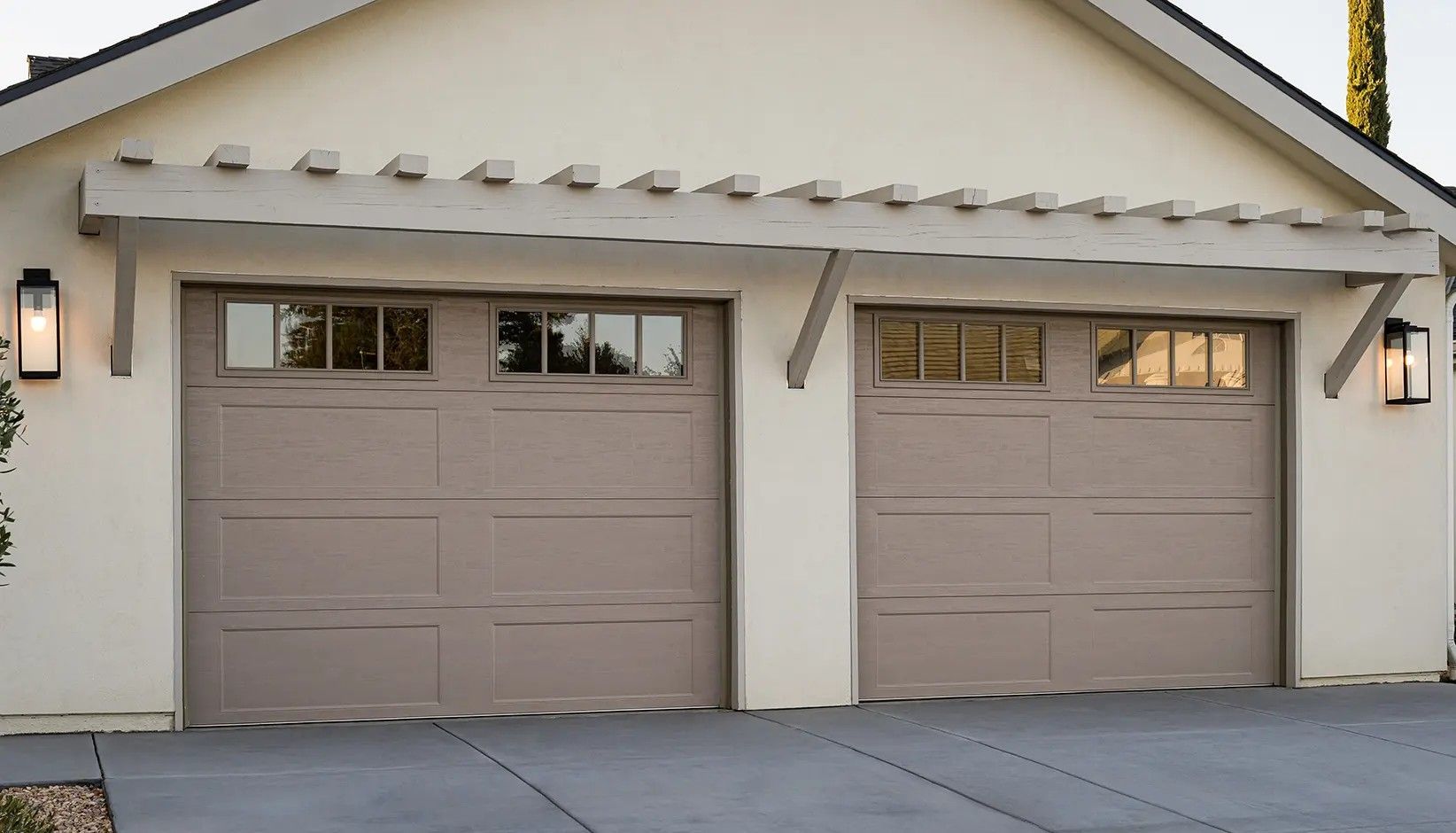 Two taupe, rectangular garage doors with windows on top, featuring a rustic wooden pergola overhead and exterior lanterns.