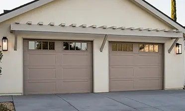 A two-car garage with taupe doors, windows at the top, and a decorative wooden trellis above against a cream wall.