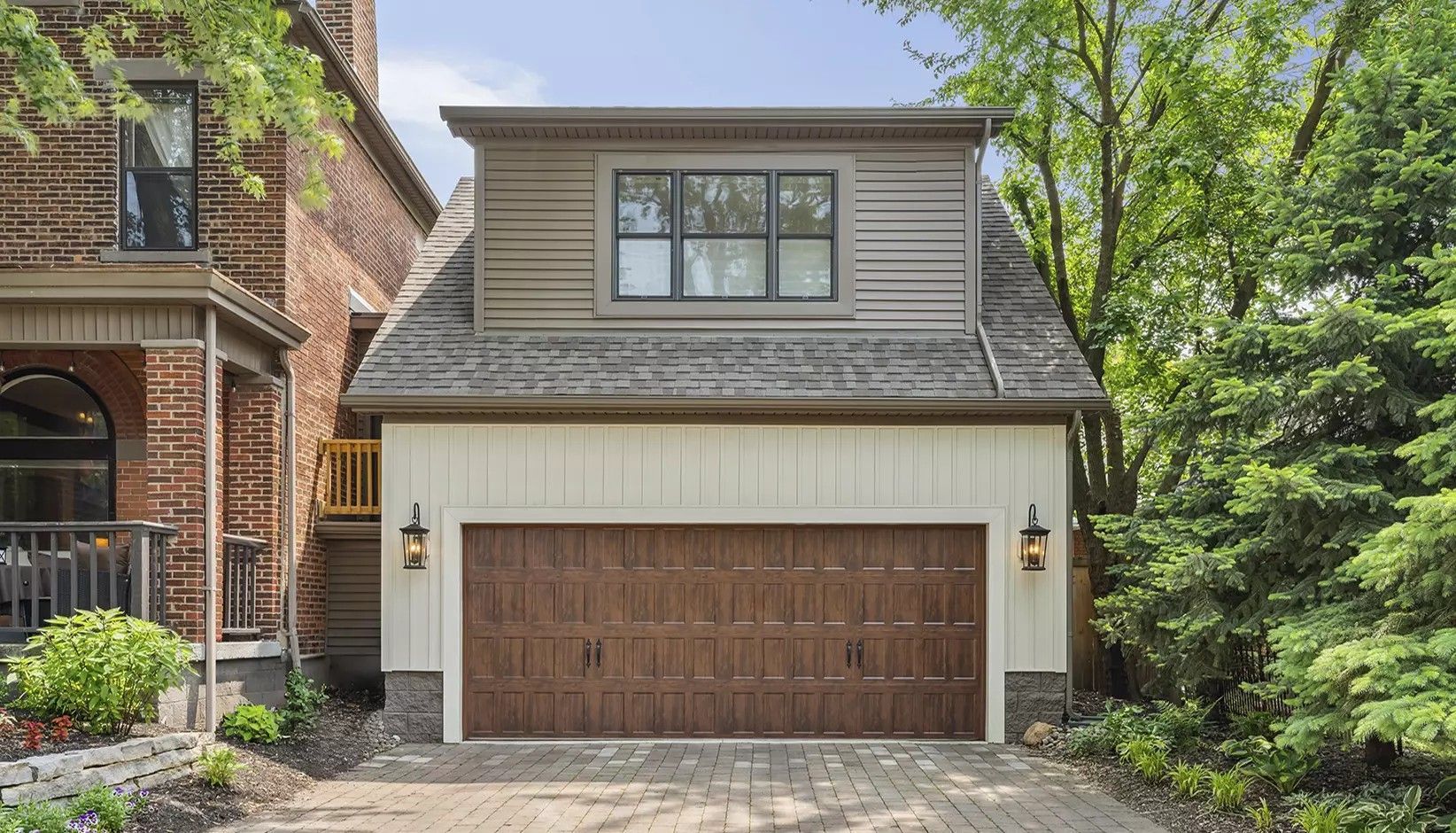 A two-story garage with a dark brown door, cream board-and-batten siding, a dormer window, and stone foundation.