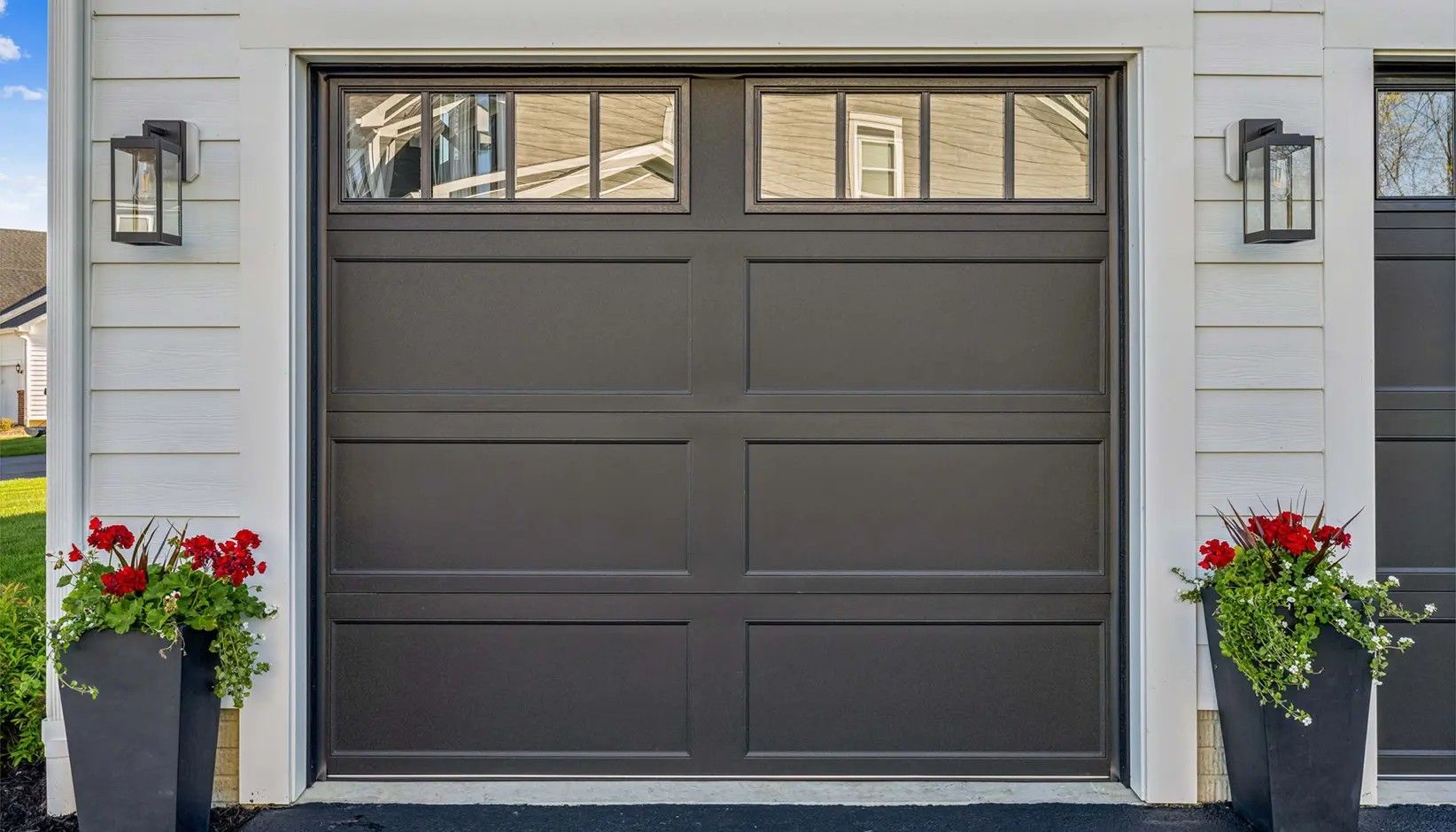 A dark brown, single-car garage door with upper windows, flanked by two tall black planters filled with red flowers.