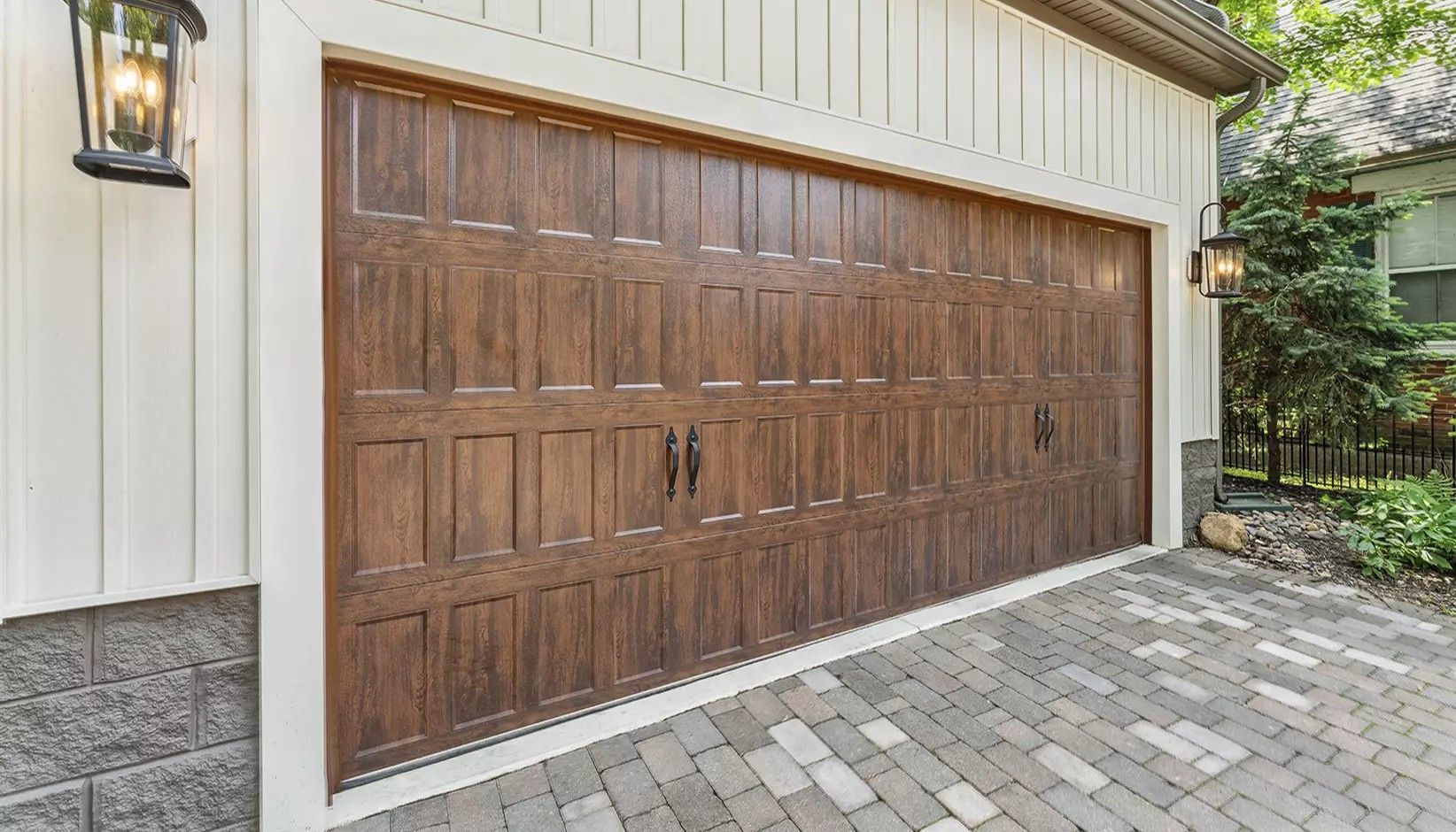 A wood-grain garage door with a rectangular panel design, set in a white-trimmed home exterior with a paver driveway.