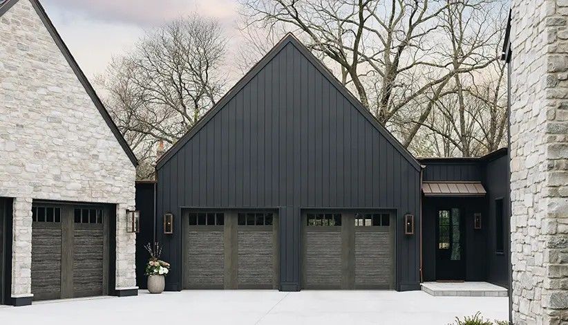 A modern home exterior with charcoal board-and-batten siding, light stone accents, and three dark wood garage doors.