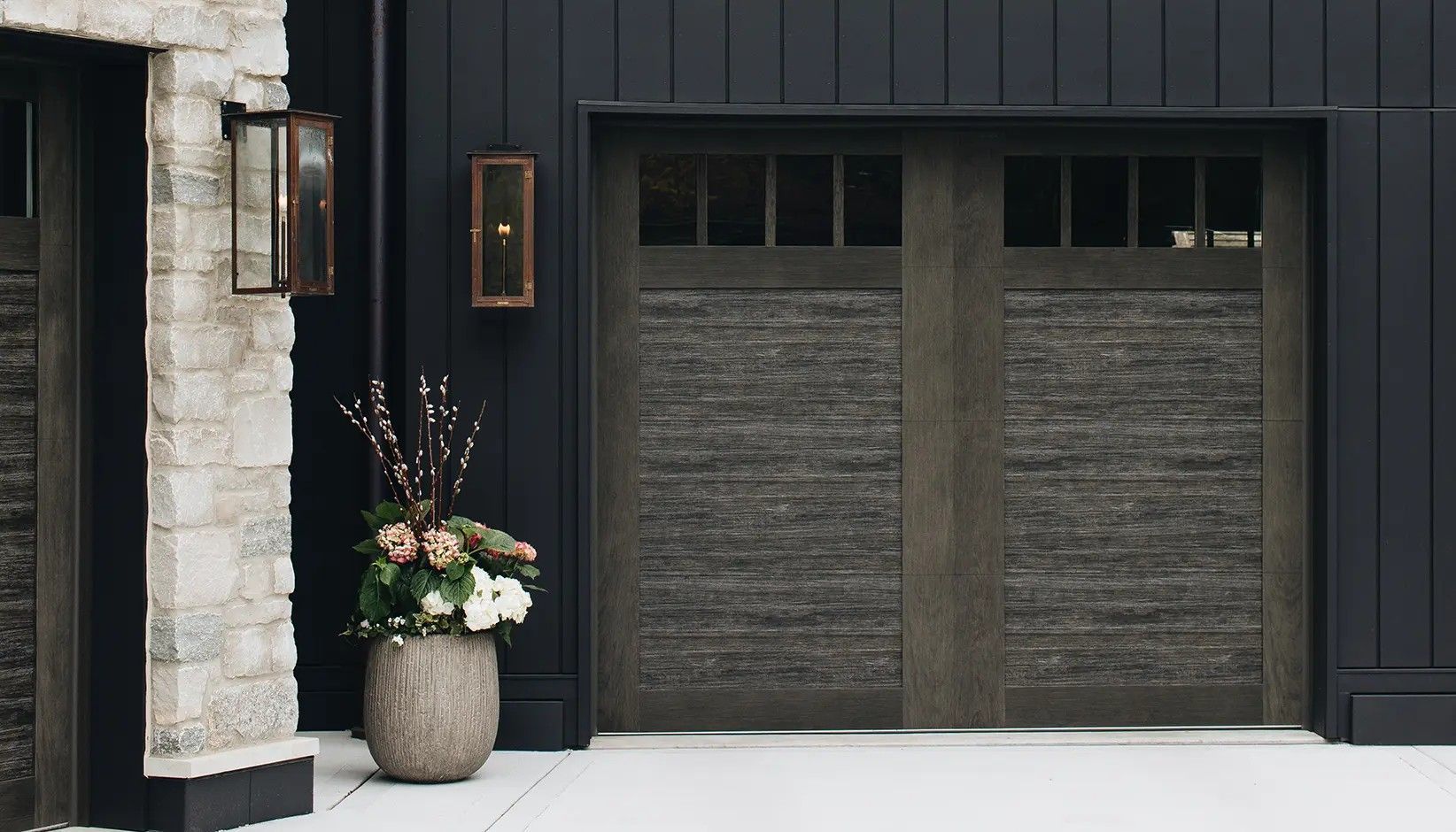 A modern garage featuring dark wood doors with rectangular windows, flanked by stone masonry and black vertical siding.
