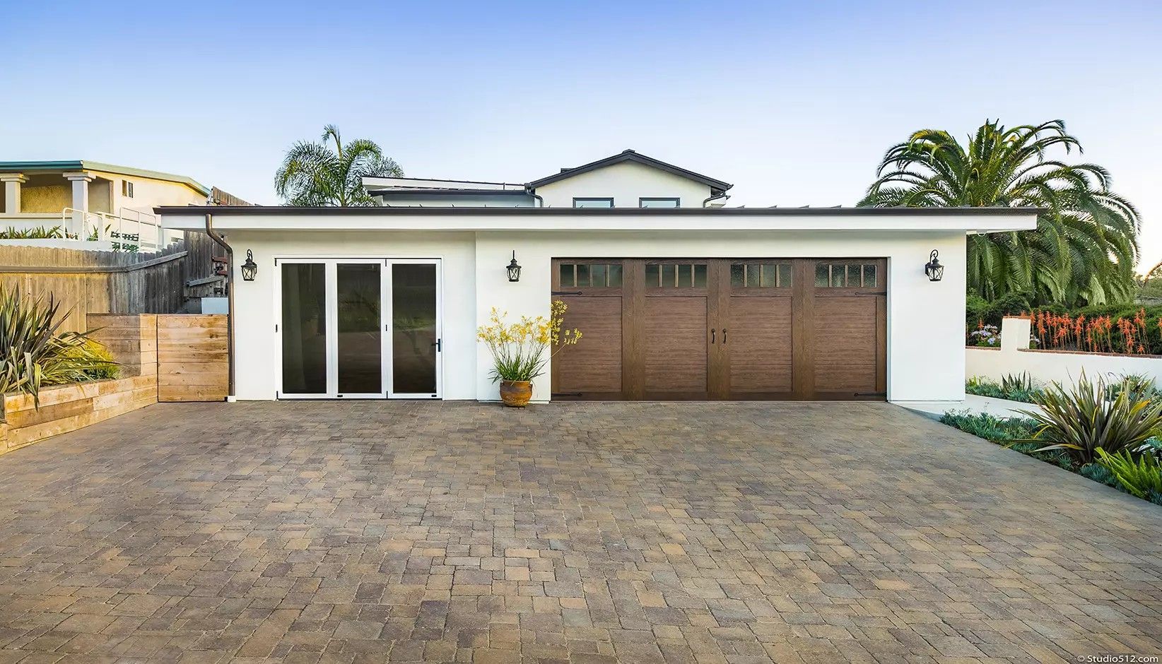 A white single-story house with a wooden two-car garage, glass patio doors, and a stone-paved driveway under a clear sky.