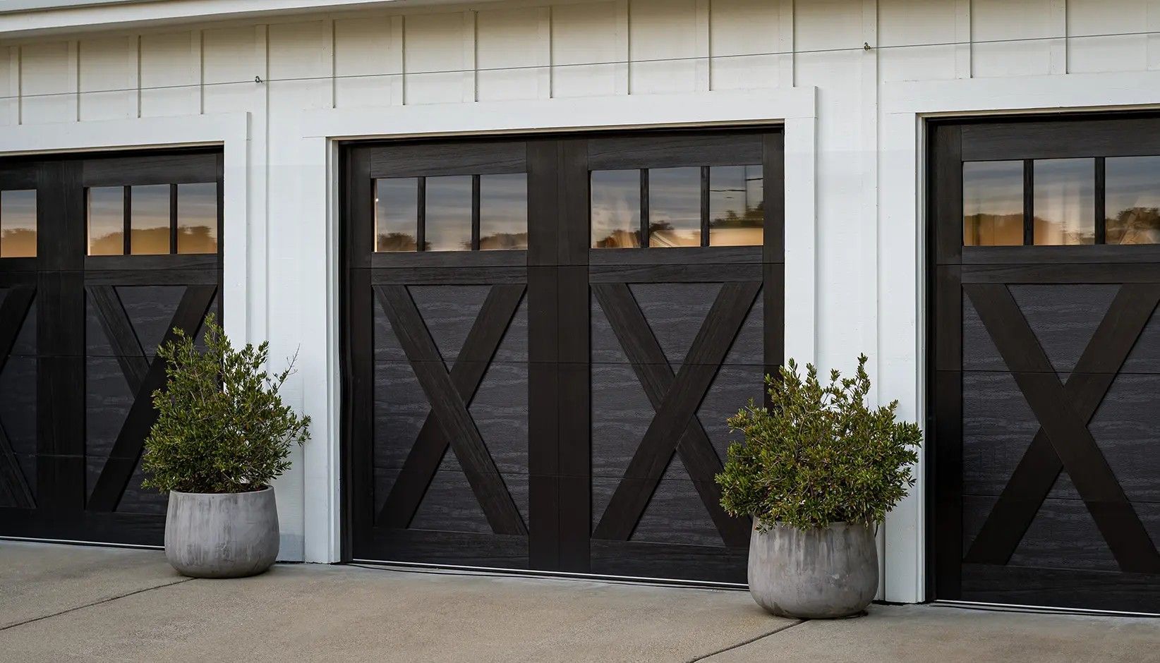 Three dark-stained wooden garage doors with cross-brace designs and upper windows, flanked by two potted green shrubs.